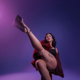 Comedian Hayley Sproull poses sitting on a stool with her leg raised.