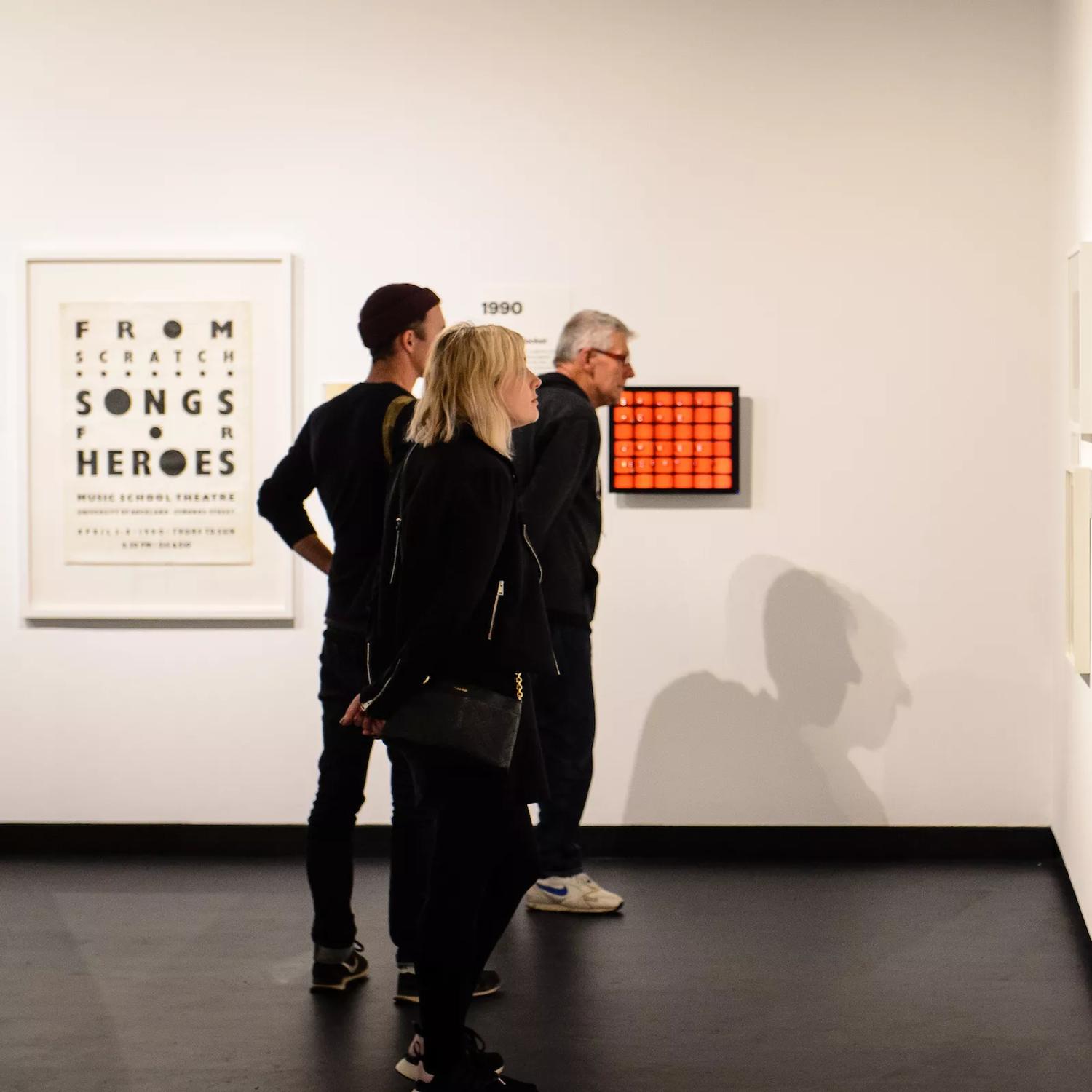 3 people wearing black, appreciating art works on the white wall inside City Gallery.