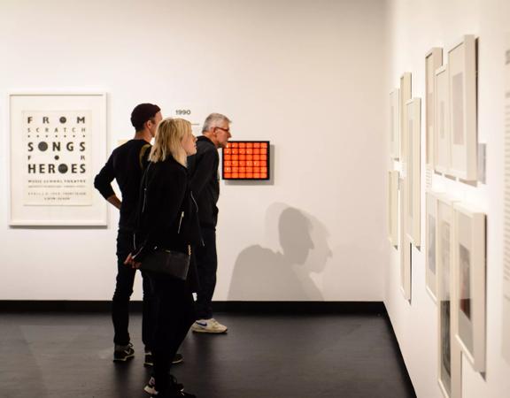 3 people wearing black, appreciating art works on the white wall inside City Gallery.