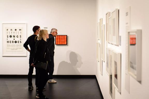 3 people wearing black, appreciating art works on the white wall inside City Gallery.