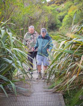 Two people walk across a flat bridge among native bush.