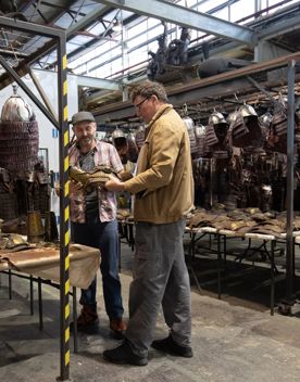 Sir Richard Taylor, founder of Wētā Workshop talks with another person. They are in a warehouse surrounded by metal helmets hanging from metal racks.