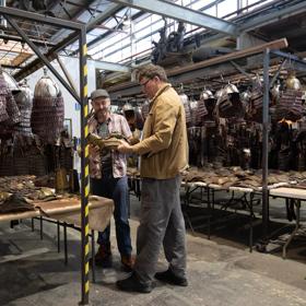 Sir Richard Taylor, founder of Wētā Workshop talks with another person. They are in a warehouse surrounded by metal helmets hanging from metal racks.