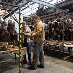 Sir Richard Taylor, founder of Wētā Workshop talks with another person. They are in a warehouse surrounded by metal helmets hanging from metal racks.