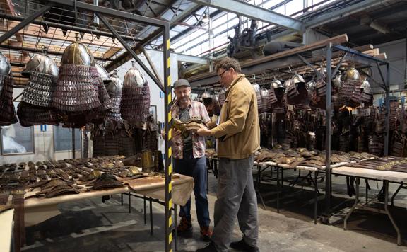 Sir Richard Taylor, founder of Wētā Workshop talks with another person. They are in a warehouse surrounded by metal helmets hanging from metal racks.