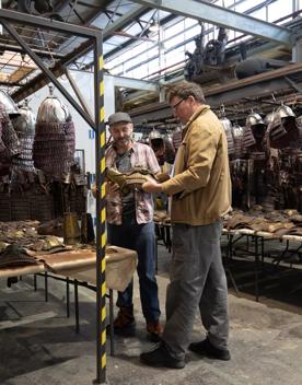 Sir Richard Taylor, founder of Wētā Workshop talks with another person. They are in a warehouse surrounded by metal helmets hanging from metal racks.