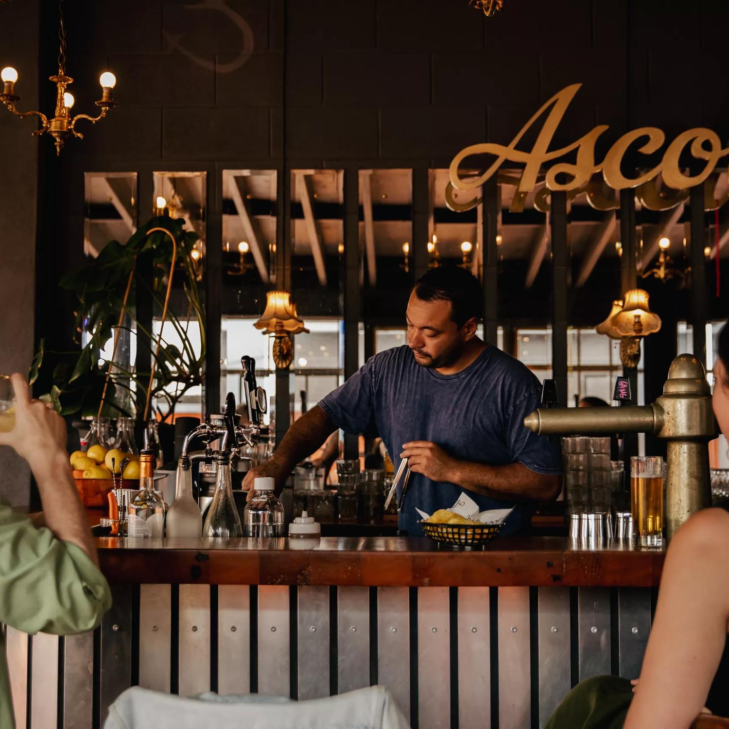 Two people sitting up at the bar at Ascot, catching up for a drink whilst being served by a bartender.