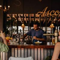 Two people sitting up at the bar at Ascot, catching up for a drink whilst being served by a bartender.