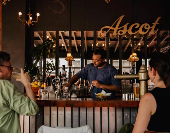 Two people sitting up at the bar at Ascot, catching up for a drink whilst being served by a bartender.