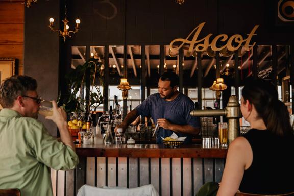 Two people sitting up at the bar at Ascot, catching up for a drink whilst being served by a bartender.