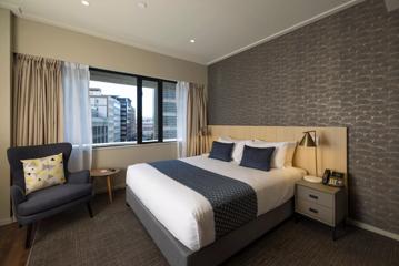 A hotel room with a queen-sized bed, grey wallpaper and carpet, and an armchair in the corner by the window.