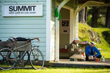 Two people are sitting on the ground with crossed legs, chatting and sharing tea. They are beside a small hut with a sign that reads "Summit height above sea level 1141 feet" on the Remutaka Cycle Trail.