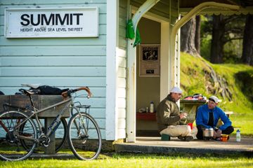 Two people are sitting on the ground with crossed legs, chatting and sharing tea. They are beside a small hut with a sign that reads "Summit height above sea level 1141 feet" on the Remutaka Cycle Trail.