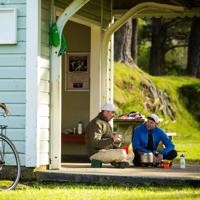Two people are sitting on the ground with crossed legs, chatting and sharing tea. They are beside a small hut with a sign that reads "Summit height above sea level 1141 feet" on the Remutaka Cycle Trail. 