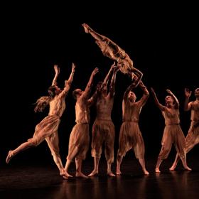 A group of dancers throw another dancer in the air. They are all dressed in brown, with a black background.