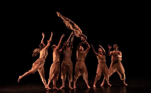 A group of dancers throw another dancer in the air. They are all dressed in brown, with a black background.