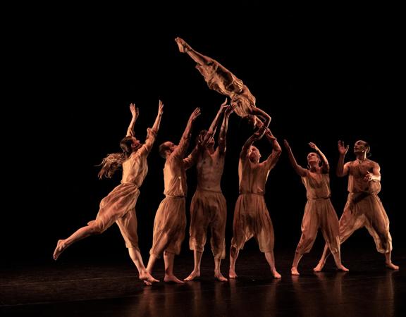 A group of dancers throw another dancer in the air. They are all dressed in brown, with a black background.