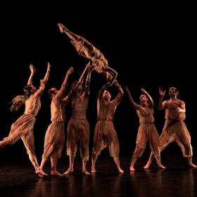 A group of dancers throw another dancer in the air. They are all dressed in brown, with a black background.