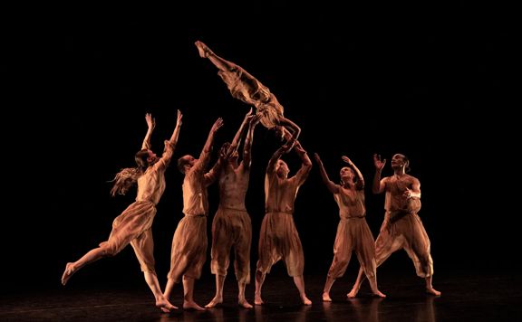 A group of dancers throw another dancer in the air. They are all dressed in brown, with a black background.