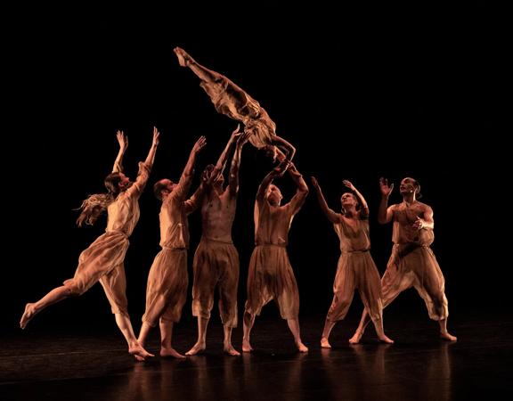 A group of dancers throw another dancer in the air. They are all dressed in brown, with a black background.