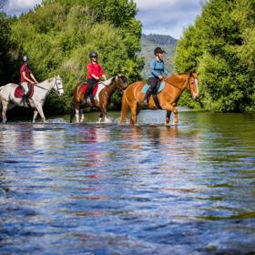 Three people cross a shallow river on horesback.