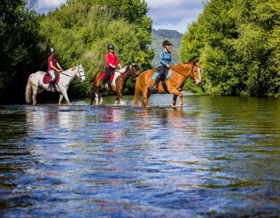 Three people cross a shallow river on horesback.