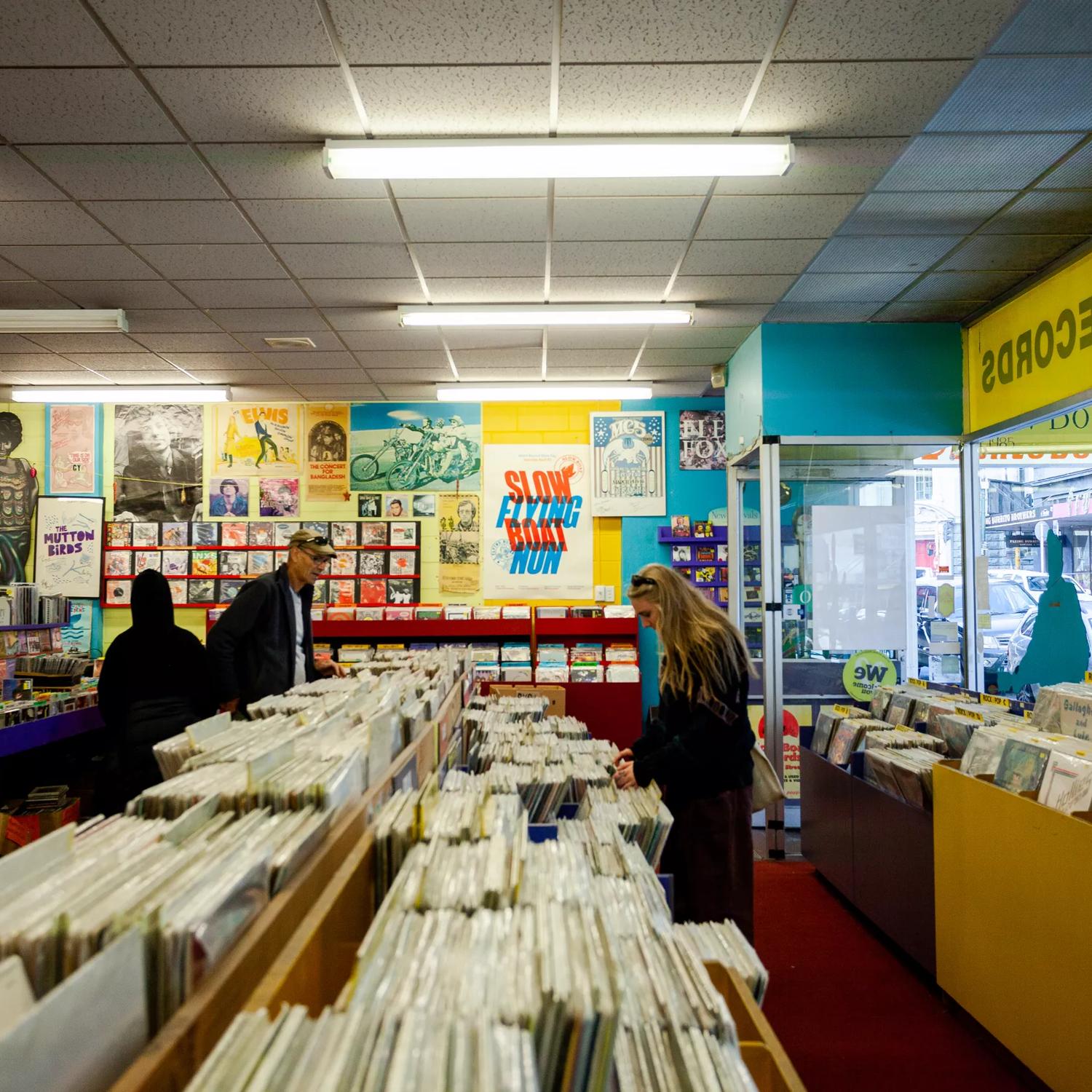 The interior of Slow Boat Records, a record store located on Cuba Street in Te Aro, Wellington. The Blue and yellow walls are covered in posters and three people browse the store's selection.