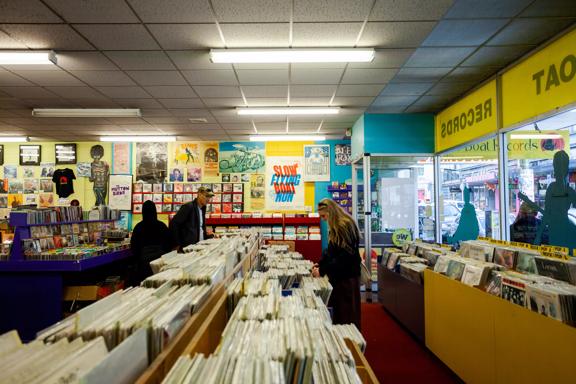 The interior of Slow Boat Records, a record store located on Cuba Street in Te Aro, Wellington. The Blue and yellow walls are covered in posters and three people browse the store's selection.