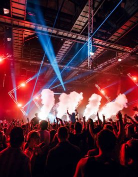 A packed crowd inside a dark venue lit up with red and blue stage, with smoke clouds coming from the stage.