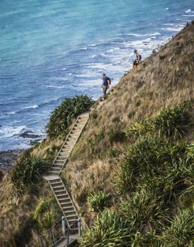 Two people walking down a wooden staircase perched on the edge of a hill, with the ocean down below.