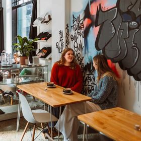 Two friends enjoy coffee in a sunny café with graffitied walls.
