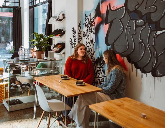 Two friends enjoy coffee in a sunny café with graffitied walls.