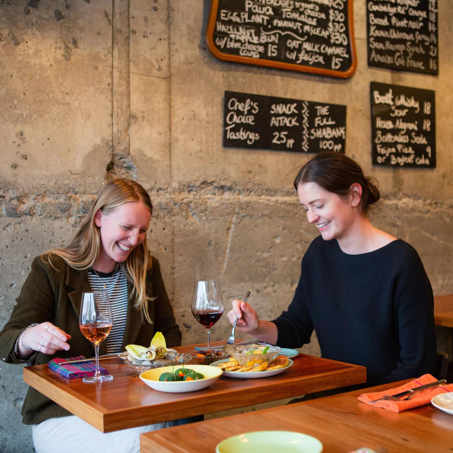 Two friends are sharing wine and snacks at GRAZE wine bar in Kelburn, Wellington. The concrete wall behind them has four chalkboards displaying the food and drink menus.