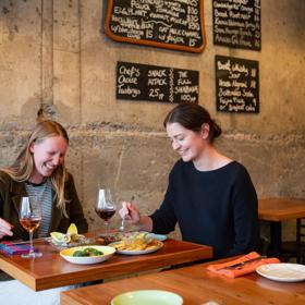 Two friends are sharing wine and snacks at GRAZE wine bar in Kelburn, Wellington. The concrete wall behind them has four chalkboards displaying the food and drink menus.