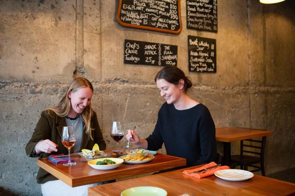 Two friends are sharing wine and snacks at GRAZE wine bar in Kelburn, Wellington. The concrete wall behind them has four chalkboards displaying the food and drink menus.