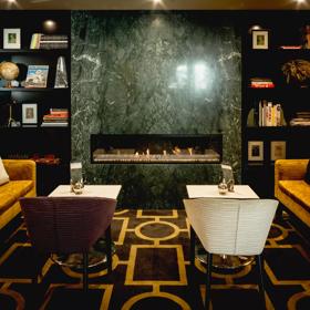 A seating area inside the Bolton Hotel with two small couches, two small white square tables with one chair each and a book shelves and a dark grey marble fireplace in the background. 