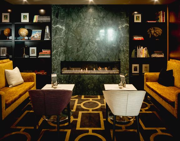 A seating area inside the Bolton Hotel with two small couches, two small white square tables with one chair each and a book shelves and a dark grey marble fireplace in the background.