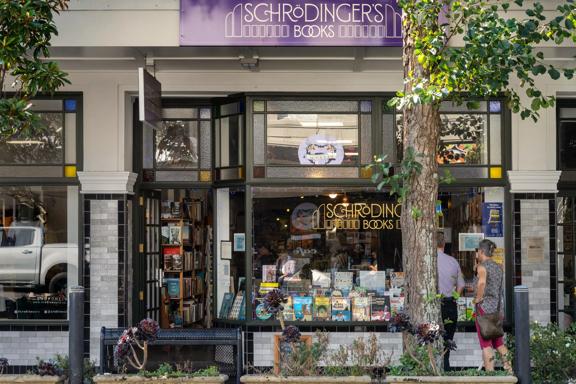 The exterior of Schrodingers books, with a vintage feel and a tree outside, as well as their purple sign above the shop. Books are displayed in the bay window.