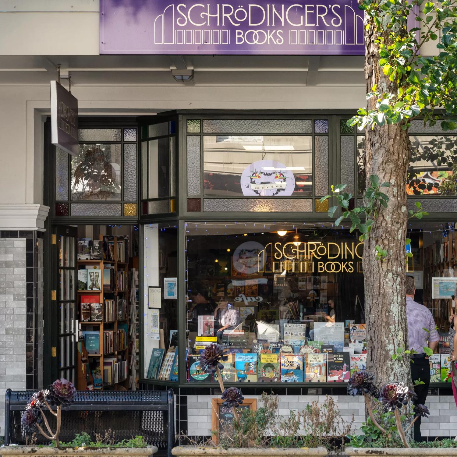 The exterior of Schrodingers books, with a vintage feel and a tree outside, as well as their purple sign above the shop. Books are displayed in the bay window.