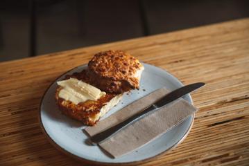 A cheese scone, sliced horizontally and buttered on a plate with a napkin and a butter knife at Swimsuit Coffee.