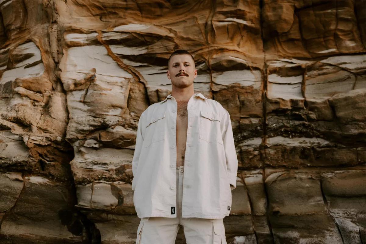 Singer-songwriter Ziggy Alberts stands in front of a rock wall with his hands in his pockets.
