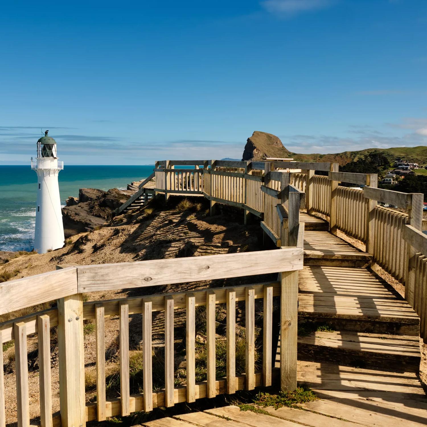 Wooden walkway leading to the Castlepoint lighthouse with hills in background and blue ocean.