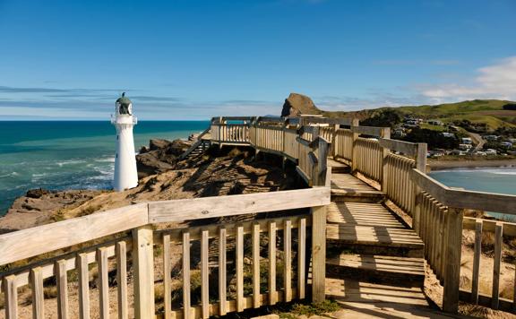 Wooden walkway leading to the Castlepoint lighthouse with hills in background and blue ocean.