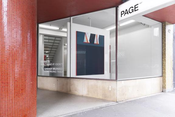 View of the outside of the Page Galleries  building from the footpath. A thick round red tiled pillar is to the left of the image. The front window of the gallery is sparse apart from one large painting. 