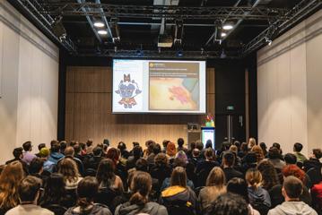Delegates at the New Zealand Game Developers Conference sit in a meeting room and look at a projector screen.