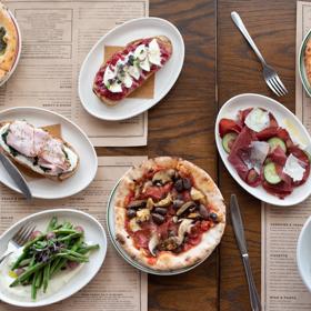 Looking down at various dishes served at Ombra on the wooden table, including pizzas, salads and Toast.