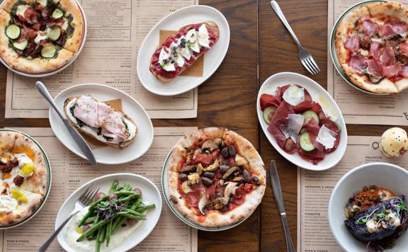 Looking down at various dishes served at Ombra on the wooden table, including pizzas, salads and Toast.