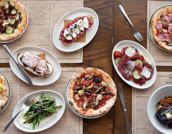 Looking down at various dishes served at Ombra on the wooden table, including pizzas, salads and Toast.