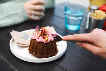 A hand using a fork to cut into a mini-cake dessert topped with pink icing on a small white plate at Maranui Café.