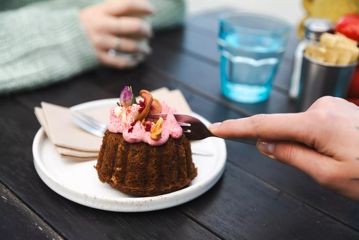 A hand using a fork to cut into a mini-cake dessert topped with pink icing on a small white plate at Maranui Café.